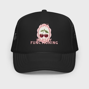 May include: A black trucker hat featuring a graphic of two red cherries with green leaves inside a light pink scalloped frame topped with a bow. Below the frame, the text BARELY FUNCTIONING is printed in light pink capital letters.