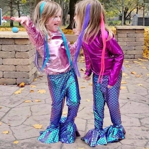 Puede incluir: Dos ni&ntilde;os vestidos con disfraces de sirena. Llevan tops brillantes y met&aacute;licos en rosa y magenta, combinados con pantalones con estampado de escamas azules y moradas con bajos acampanados. Sus cabellos est&aacute;n peinados con extensiones coloridas, y est&aacute;n de pie en un patio de piedra.