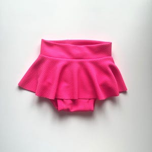 May include: A bright pink baby skirt with a textured fabric. The skirt has a high waistband and a flared, ruffled design. The garment is designed to be worn as a bottom.