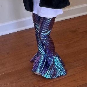 Puede incluir: Un ni&ntilde;o con pantalones acampanados con escamas de sirena en tonos azules, morados y turquesas. Los pantalones tienen una textura escamosa y un bajo acampanado. El ni&ntilde;o tambi&eacute;n lleva una camiseta blanca y una chaqueta negra.