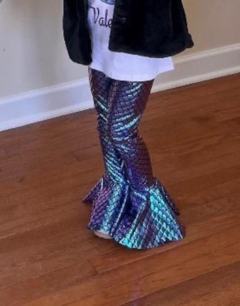 Puede incluir: Un ni&ntilde;o con pantalones acampanados con escamas de sirena en tonos azules, morados y turquesas. Los pantalones tienen una textura escamosa y un bajo acampanado. El ni&ntilde;o tambi&eacute;n lleva una camiseta blanca y una chaqueta negra.
