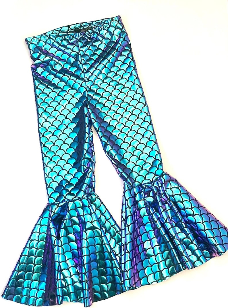 Puede incluir: Leggings azules y morados con escamas de sirena y bajos acampanados. Los leggings tienen un acabado brillante e iridiscente y un patr&oacute;n de escamas repetido. La tela parece ser un material el&aacute;stico, adecuado para disfraces o uso diario.