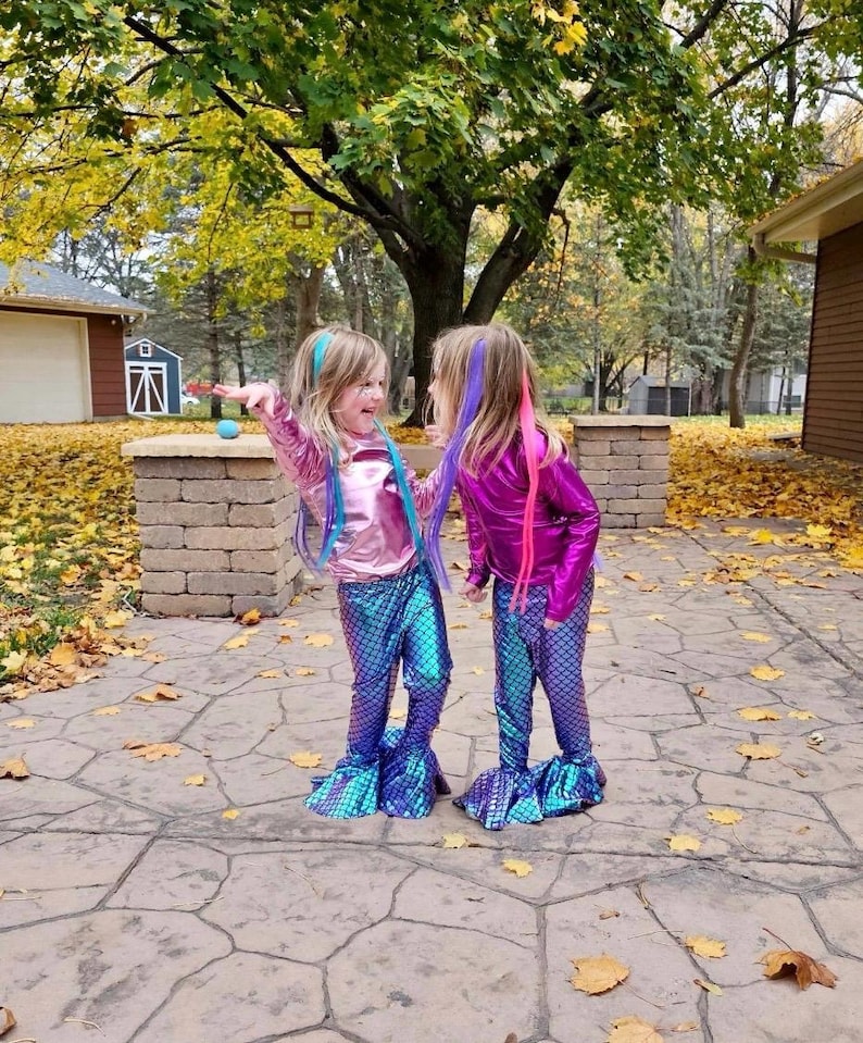 Puede incluir: Dos ni&ntilde;os vestidos con disfraces de sirena. Llevan tops brillantes y met&aacute;licos en rosa y magenta, combinados con pantalones con estampado de escamas azules y moradas con bajos acampanados. Sus cabellos est&aacute;n peinados con extensiones coloridas, y est&aacute;n de pie en un patio de piedra.
