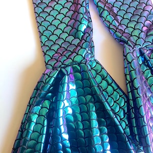 Puede incluir: Leggings iridiscentes con escamas de sirena en tonos turquesa, morado y azul. Los leggings presentan un bajo acampanado, creando un efecto de cola de sirena. La tela tiene una calidad brillante y reflectante, y el patr&oacute;n de escamas est&aacute; delineado en negro.