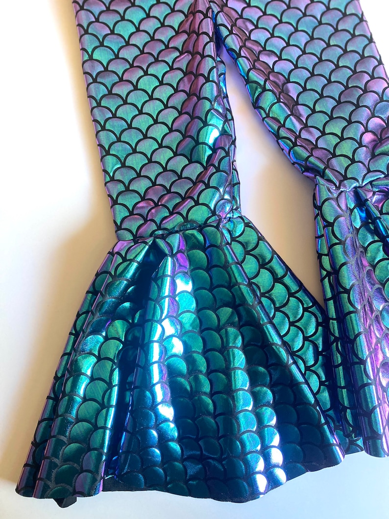 Puede incluir: Leggings iridiscentes con escamas de sirena en tonos turquesa, morado y azul. Los leggings presentan un bajo acampanado, creando un efecto de cola de sirena. La tela tiene una calidad brillante y reflectante, y el patr&oacute;n de escamas est&aacute; delineado en negro.