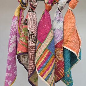 May include: A row of colorful, quilted scarves hanging from a metal rod. The scarves feature a variety of patterns, including floral, geometric, and patchwork designs. The color palette includes pink, orange, teal, and purple.