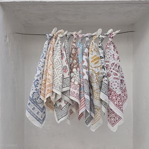 May include: A collection of patterned fabric squares, likely bandanas or small scarves, hang from a wire. The fabrics feature various designs in colors like blue, red, yellow, and brown. They are displayed against a white wall with a pink accent.