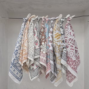 May include: A collection of square, patterned fabric cloths in various colors, including blue, yellow, brown, and red, are displayed hanging from a wire. Each cloth features a unique floral or geometric design, with white borders and knotted tops.