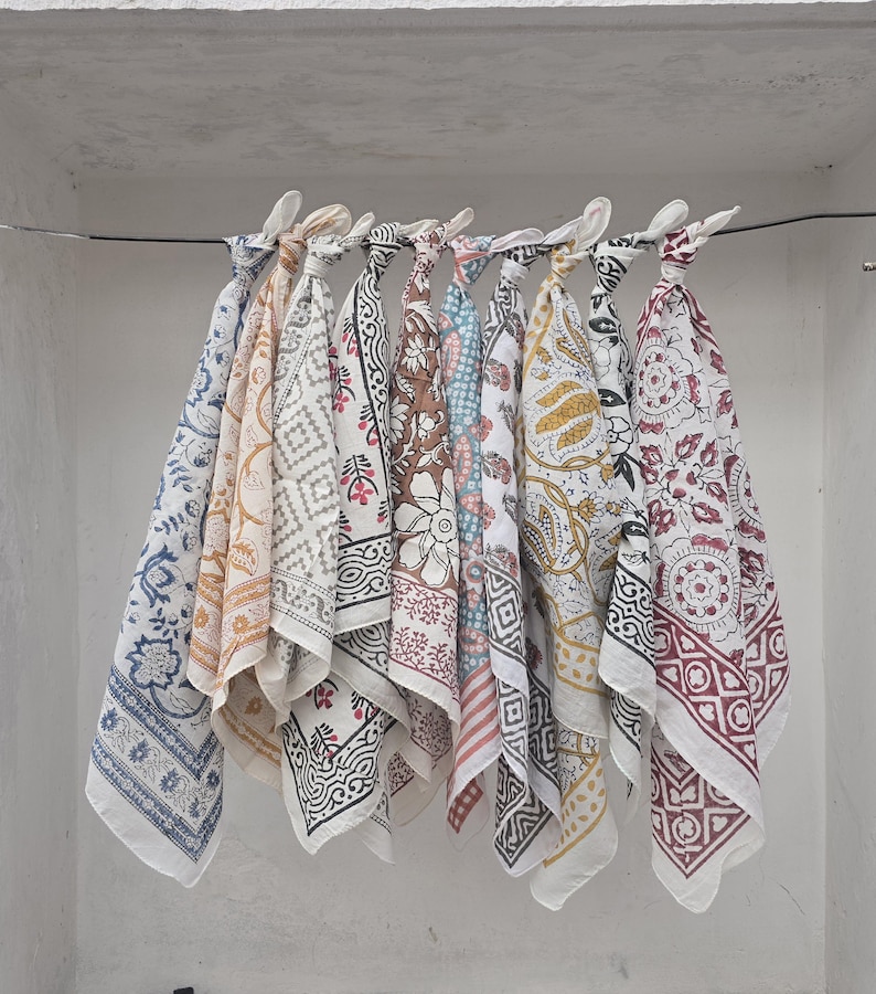 May include: A collection of square, patterned fabric cloths in various colors, including blue, yellow, brown, and red, are displayed hanging from a wire. Each cloth features a unique floral or geometric design, with white borders and knotted tops.