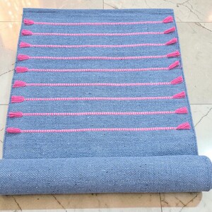 May include: A rolled-up blue woven rug with horizontal pink stripes and pink tassels. The rug is made of a textured material and is partially rolled up, revealing the underside.