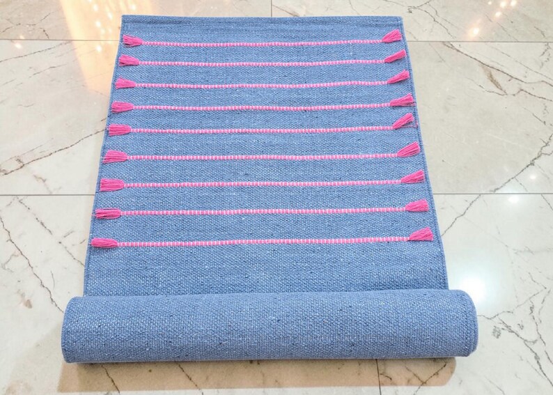 May include: A rolled-up blue woven rug with horizontal pink stripes and pink tassels. The rug is made of a textured material and is partially rolled up, revealing the underside.