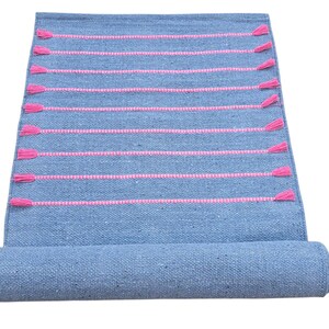 May include: A rolled-up blue woven rug with horizontal pink stripes and pink tassels. The rug is made of a textured material and is partially rolled up, revealing the underside. The tassels are evenly spaced along the edges.