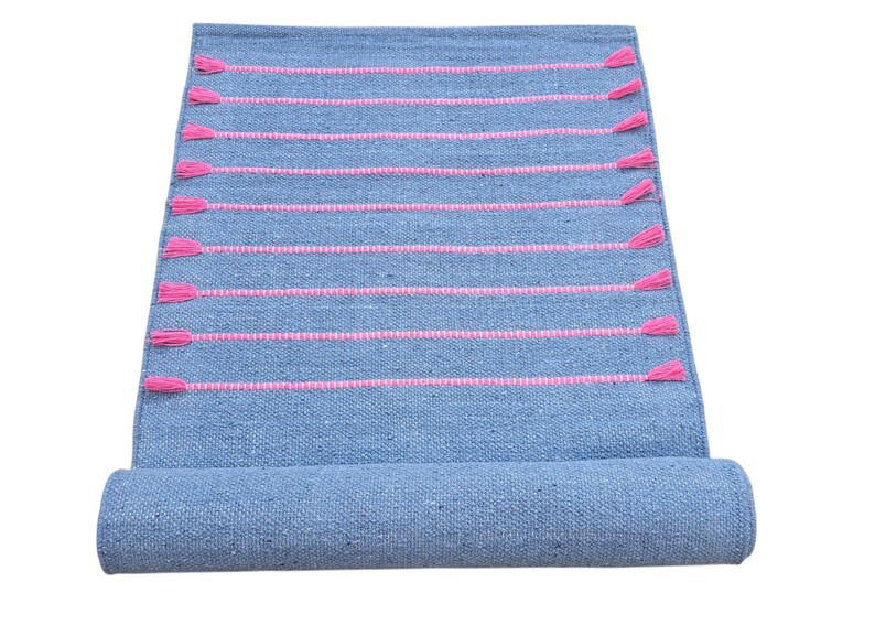 May include: A rolled-up blue woven rug with horizontal pink stripes and pink tassels. The rug is made of a textured material and is partially rolled up, revealing the underside. The tassels are evenly spaced along the edges.