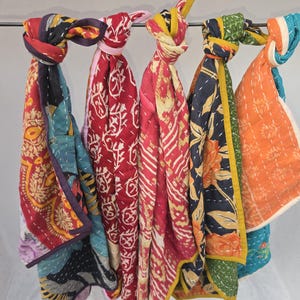 Op de afbeelding: Een verzameling kleurrijke, bedrukte sjaals die aan een metalen stang hangen. De sjaals hebben verschillende ontwerpen, waaronder bloemen- en geometrische patronen, in tinten rood, blauw, oranje en geel. Elke sjaal is bovenaan geknoopt.