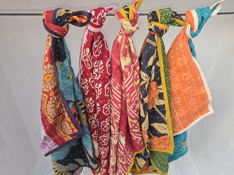 Op de afbeelding: Een verzameling kleurrijke, bedrukte sjaals die aan een metalen stang hangen. De sjaals hebben verschillende ontwerpen, waaronder bloemen- en geometrische patronen, in tinten rood, blauw, oranje en geel. Elke sjaal is bovenaan geknoopt.