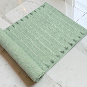 May include: A rectangular, light green rug with vertical white stripes and small tassels along the edges. The rug is partially rolled up, revealing its textured weave. The rug is placed on a marble tile surface.
