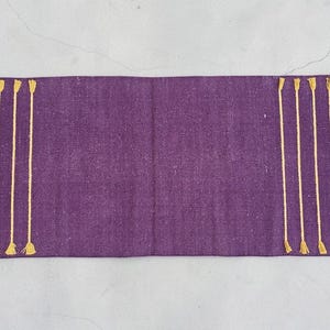 May include: A rectangular, purple table runner with vertical gold-colored tassel accents on each end. The runner is made of a textured fabric and is displayed on a light gray surface.