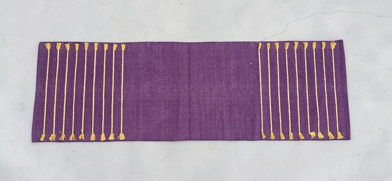May include: A rectangular, purple table runner with vertical gold-colored tassel accents on each end. The runner is made of a textured fabric and is displayed on a light gray surface.