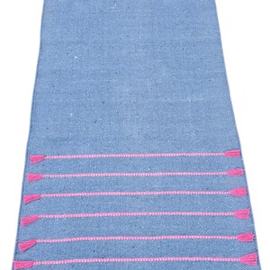 May include: A rectangular blue woven rug with horizontal pink stripes and pink tassels at each end. The rug is made of a textured material and is suitable for use as a runner.