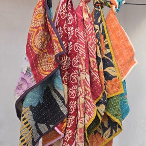 Op de afbeelding: Een verzameling kleurrijke, gewatteerde stoffen vierkanten, elk met unieke patronen en ontwerpen. De stoffen zijn in tinten rood, oranje, blauw en zwart, met bloemen- en geometrische motieven. De vierkanten hangen aan een stang.