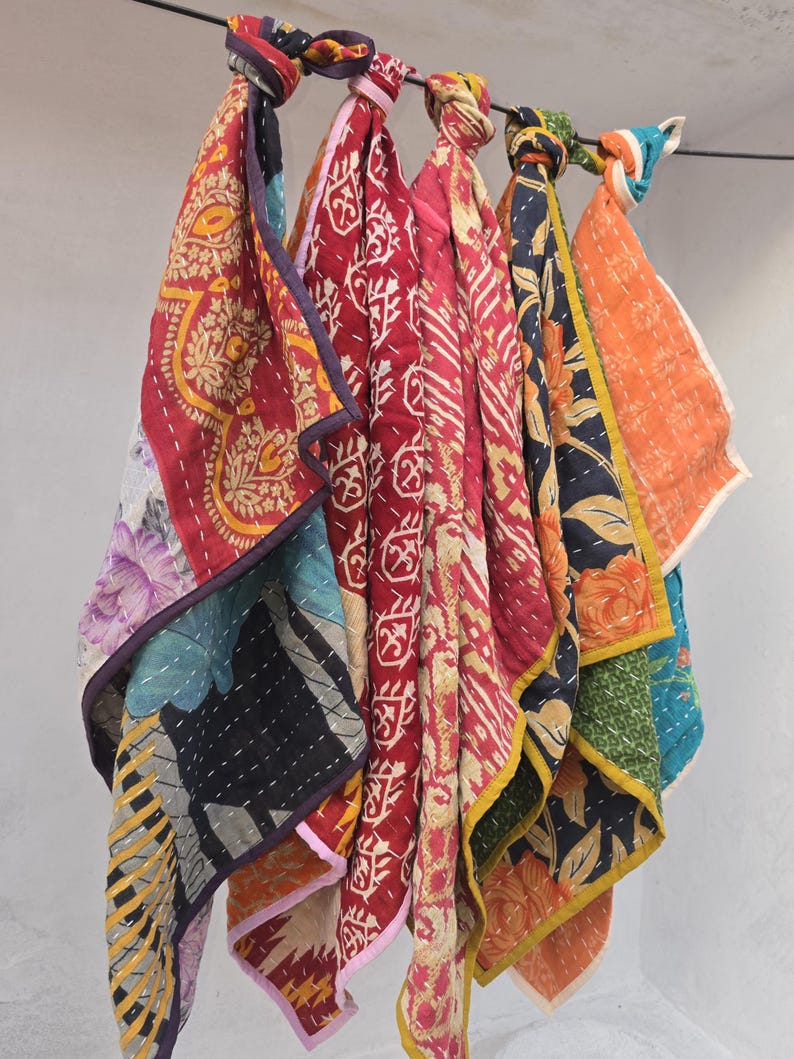 Op de afbeelding: Een verzameling kleurrijke, gewatteerde stoffen vierkanten, elk met unieke patronen en ontwerpen. De stoffen zijn in tinten rood, oranje, blauw en zwart, met bloemen- en geometrische motieven. De vierkanten hangen aan een stang.