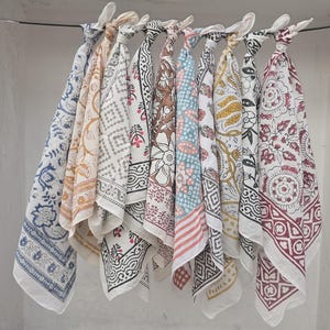 May include: A collection of patterned fabric squares, likely bandanas or scarves, hang from a metal rod. The designs include floral, geometric, and paisley patterns in various colors like blue, red, brown, and yellow. The fabric is lightweight and appears to be cotton.