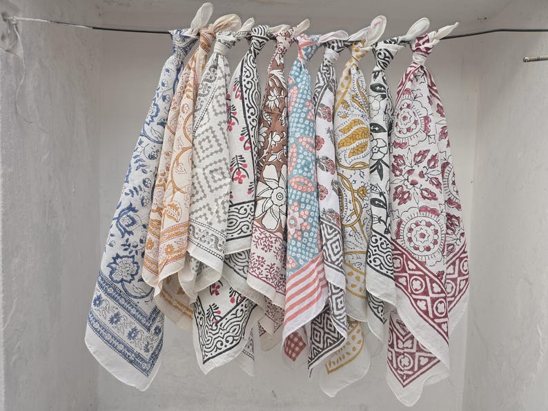 May include: A collection of patterned fabric squares, likely bandanas or scarves, hang from a metal rod. The designs include floral, geometric, and paisley patterns in various colors like blue, red, brown, and yellow. The fabric is lightweight and appears to be cotton.