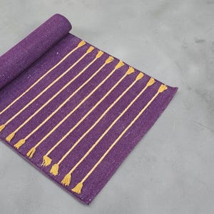 May include: A rolled-up purple rug with vertical gold stripes and tassels. The rug is made of a woven material and is partially unrolled, displaying the striped pattern. The tassels are at the bottom of the unrolled section.