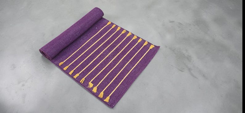 May include: A rolled-up purple rug with vertical gold stripes and tassels. The rug is made of a woven material and is partially unrolled, displaying the striped pattern. The tassels are at the bottom of the unrolled section.