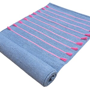 May include: A rolled-up blue rug with pink vertical stripes and tassels. The rug is made of woven material and has a textured appearance. The pink stripes and tassels add a pop of color to the otherwise neutral blue.