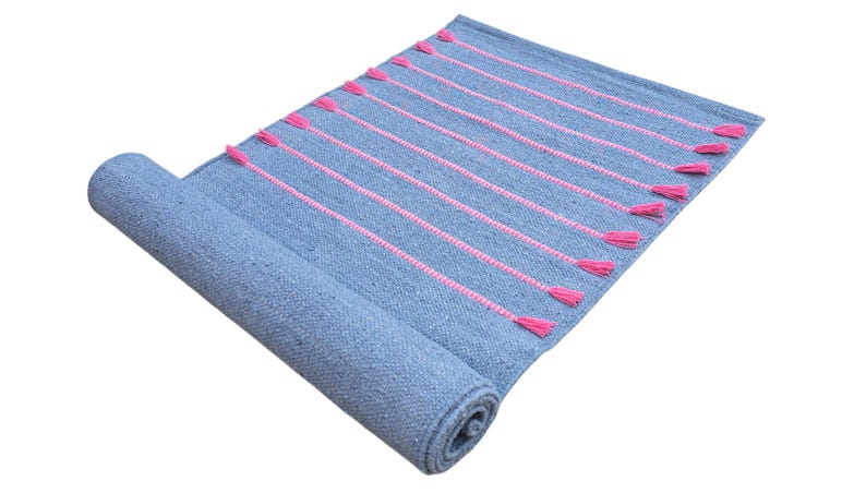 May include: A rolled-up blue rug with pink vertical stripes and tassels. The rug is made of woven material and has a textured appearance. The pink stripes and tassels add a pop of color to the otherwise neutral blue.