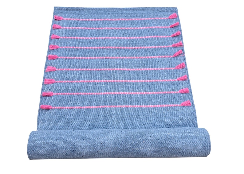 May include: A rolled-up blue woven rug with horizontal pink stripes and pink tassels. The rug is made of a textured material and is displayed against a white background.