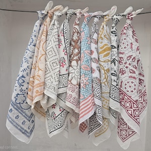 May include: A collection of patterned fabric squares, likely bandanas or scarves, hang from a rod. The designs include floral, geometric, and paisley patterns in various colors like blue, brown, pink, and yellow. The fabric is white with the designs printed on it.