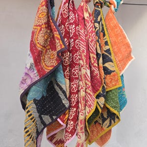 Op de afbeelding: Een verzameling kleurrijke, gewatteerde stoffen sjaals die aan een zwarte stang hangen. De sjaals hebben verschillende patronen, waaronder bloemen- en geometrische ontwerpen, in tinten rood, oranje, blauw en geel. Elke sjaal is bovenaan geknoopt.