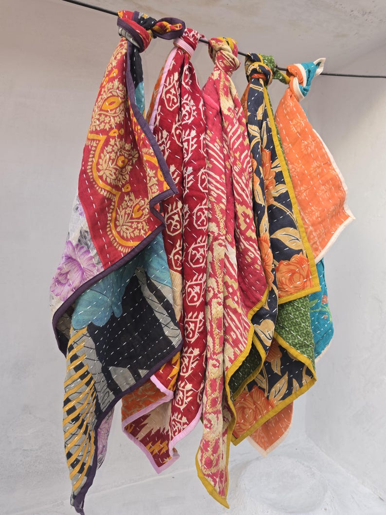Op de afbeelding: Een verzameling kleurrijke, gewatteerde stoffen sjaals die aan een zwarte stang hangen. De sjaals hebben verschillende patronen, waaronder bloemen- en geometrische ontwerpen, in tinten rood, oranje, blauw en geel. Elke sjaal is bovenaan geknoopt.