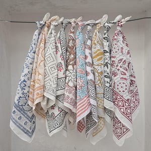 May include: A collection of nine patterned cotton bandanas in various colors, including blue, brown, and yellow, are displayed hanging from a wire. Each bandana features a unique block print design, showcasing floral and geometric patterns.