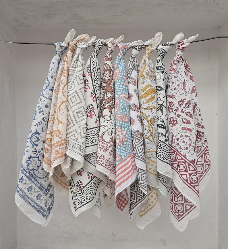 May include: A collection of nine patterned cotton bandanas in various colors, including blue, brown, and yellow, are displayed hanging from a wire. Each bandana features a unique block print design, showcasing floral and geometric patterns.