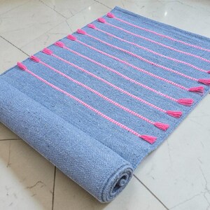 May include: A rolled-up, rectangular blue rug with pink vertical stripes and tassels. The rug is made of woven material and is partially rolled, revealing the underside. The tassels are a bright pink color.