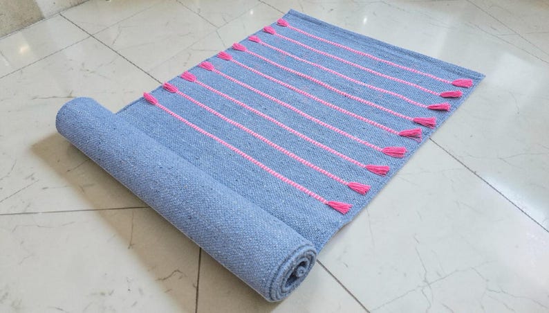 May include: A rolled-up, rectangular blue rug with pink vertical stripes and tassels. The rug is made of woven material and is partially rolled, revealing the underside. The tassels are a bright pink color.