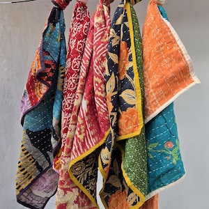 Op de afbeelding: Een verzameling kleurrijke, bedrukte stoffen sjaals die aan een metalen staaf hangen. De sjaals hebben verschillende ontwerpen, waaronder bloemen- en geometrische patronen, in tinten rood, blauw, oranje en groen. Elke sjaal is bovenaan geknoopt.