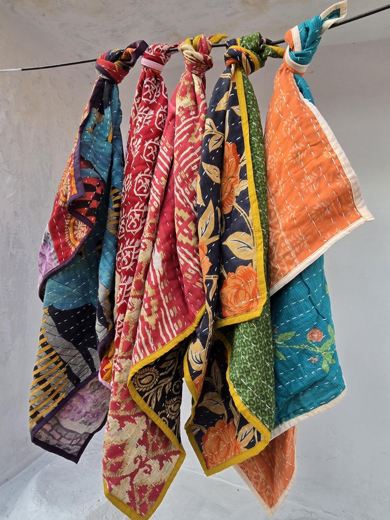 Op de afbeelding: Een verzameling kleurrijke, bedrukte stoffen sjaals die aan een metalen staaf hangen. De sjaals hebben verschillende ontwerpen, waaronder bloemen- en geometrische patronen, in tinten rood, blauw, oranje en groen. Elke sjaal is bovenaan geknoopt.