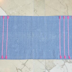 May include: A rectangular, light blue woven rug with vertical pink embroidered lines. The pink lines are evenly spaced and end in a small, triangular design. The rug is displayed on a marble floor.