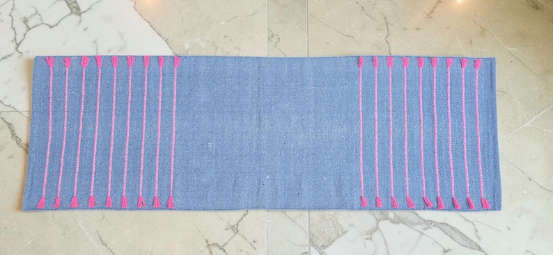 May include: A rectangular, light blue woven rug with vertical pink embroidered lines. The pink lines are evenly spaced and end in a small, triangular design. The rug is displayed on a marble floor.