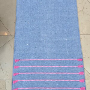 May include: A rectangular, light blue rug with horizontal pink stripes and pink tassels at the ends. The rug is made of a woven material, suitable for a hallway or entryway.