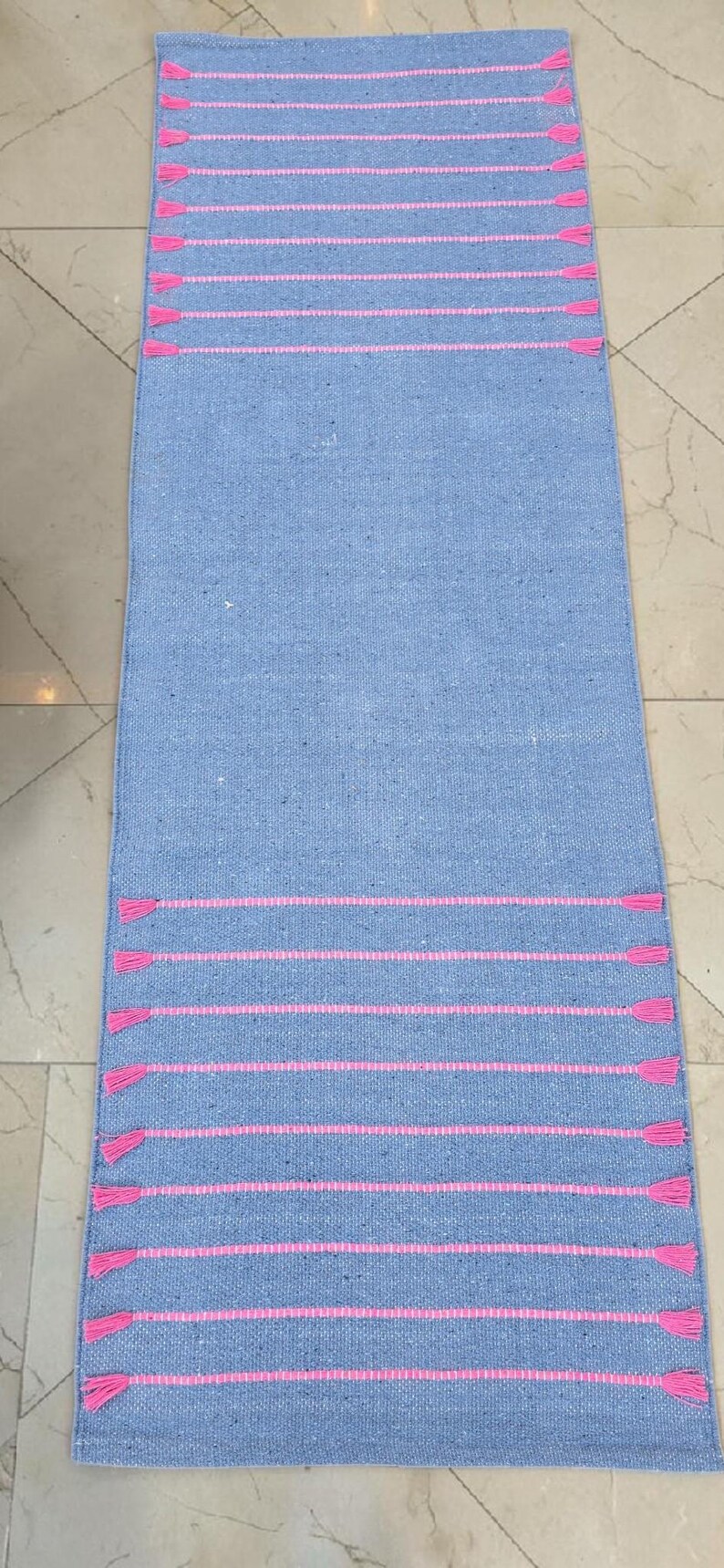May include: A rectangular, light blue rug with horizontal pink stripes and pink tassels at the ends. The rug is made of a woven material, suitable for a hallway or entryway.