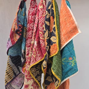 Op de afbeelding: Een verzameling van vijf kleurrijke, bedrukte sjaals hangend aan een zwarte draad. De sjaals hebben verschillende ontwerpen, waaronder bloemen- en geometrische patronen, in tinten rood, blauw, oranje en groen. Elke sjaal is bovenaan geknoopt.