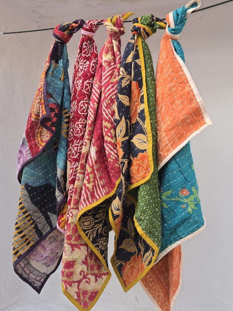 Op de afbeelding: Een verzameling van vijf kleurrijke, bedrukte sjaals hangend aan een zwarte draad. De sjaals hebben verschillende ontwerpen, waaronder bloemen- en geometrische patronen, in tinten rood, blauw, oranje en groen. Elke sjaal is bovenaan geknoopt.