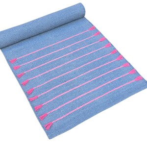 May include: A rolled-up blue rug with pink horizontal stripes and tassels. The rug is made of woven material and is displayed on a white background. The tassels are at the end of each pink stripe.