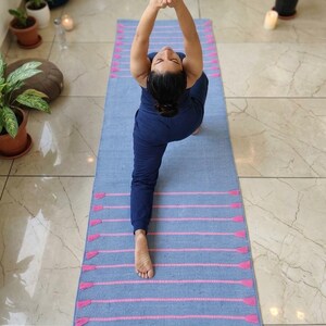 May include: A blue yoga mat with pink stripes and tassels. The mat is rectangular and is being used by someone wearing a blue outfit. The person is in a yoga pose with their hands together above their head.