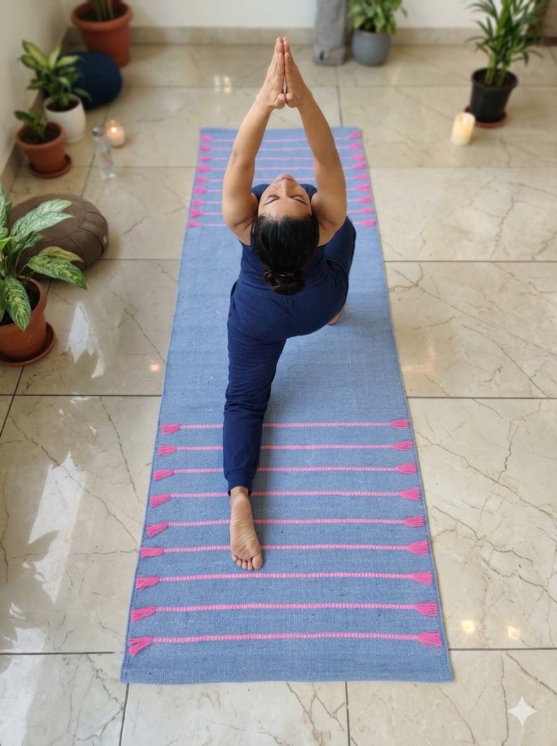May include: A blue yoga mat with pink stripes and tassels. The mat is rectangular and is being used by someone wearing a blue outfit. The person is in a yoga pose with their hands together above their head.